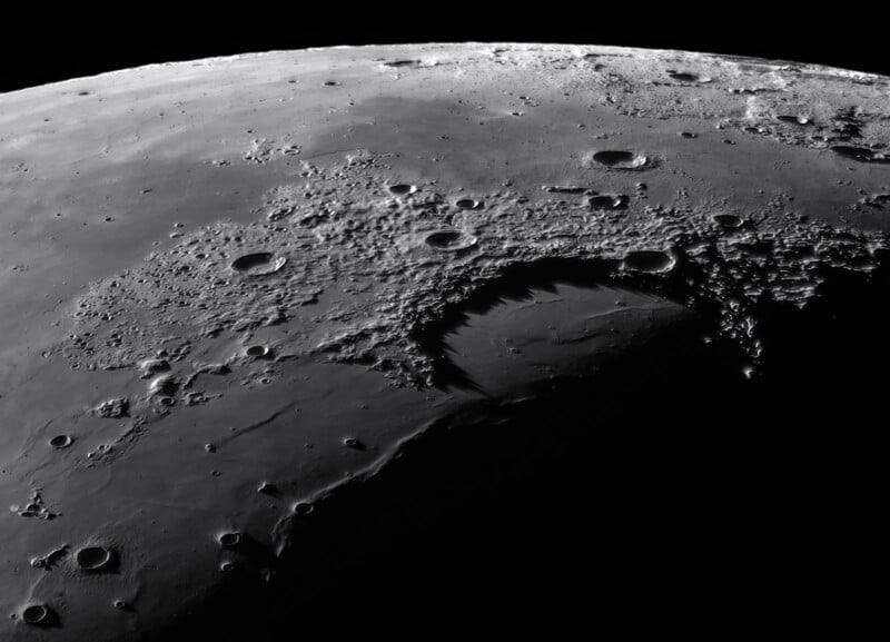 A close-up of the lunar surface showing a detailed view of numerous craters. A large shadow-filled crater is clearly visible, casting dark silhouettes against the Moon's rugged, cratered terrain. The image presents a stark and intriguing lunar landscape.