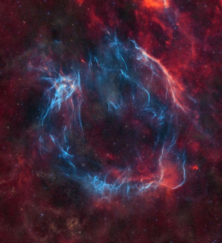 A stunning image of a cosmic scene features a ring-shaped supernova remnant. Bright blue filaments of gas and dust intertwine with fiery red and orange hues, creating an ethereal and mesmerizing spectacle against a dark background filled with stars.