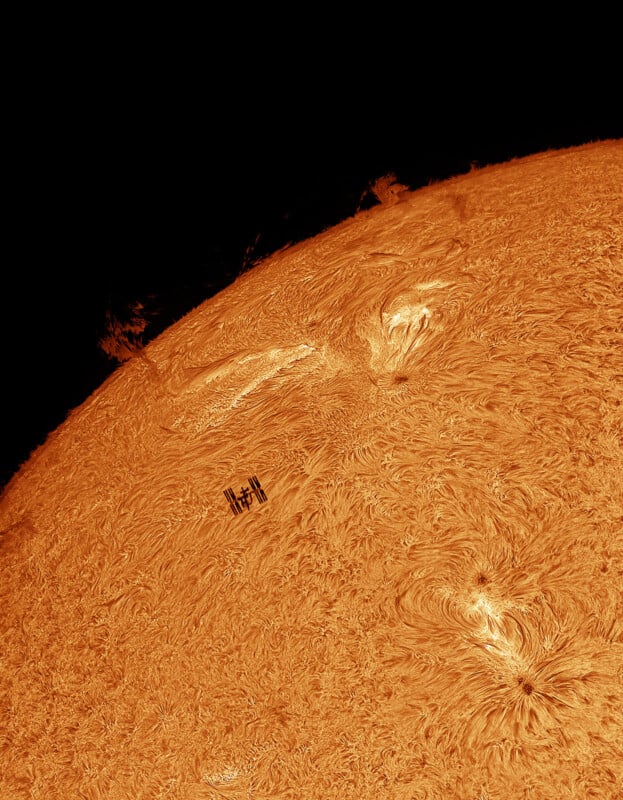 A detailed image of the Sun's surface with swirling patterns and solar prominences. The International Space Station (ISS) appears as a small silhouette against the bright background of the Sun, emphasizing its immense scale by comparison.