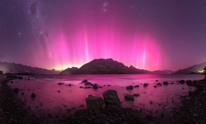 A serene lakeside landscape under a night sky illuminated by vibrant pink aurora borealis. Dark mountains are silhouetted against the brilliant display, with rocky shores framing the foreground. Soft reflections of the aurora shimmer on the surface of the calm water.