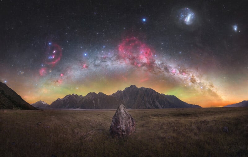 A vast night sky lit up with bright stars and colorful nebulae overlooks a mountainous landscape. In the foreground, a large, solitary rock stands in a grassy field. Snow-capped peaks and rugged cliffs rise in the distance under the vibrant sky.