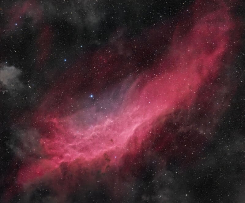 A colorful nebula with bright pink and red hues against a dark space background. Several bright stars are scattered across the image, adding contrast to the bright cloudy formation of gas and dust. The nebula appears vast and ethereal.